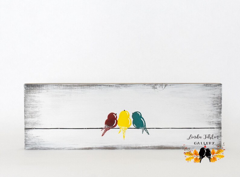 May include: A rectangular wooden sign with a distressed white finish featuring a black horizontal line with three stylized birds in maroon, yellow, and teal. The bottom right corner displays the text Linda Fehlen GALLERY with a leaf graphic.