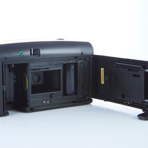 May include: A black and gold 35mm film camera with the back open, revealing the film compartment and a black film cassette. The camera has a small green light on the top.