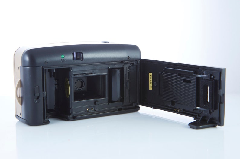 May include: A black and gold 35mm film camera with the back open, revealing the film compartment and a black film cassette. The camera has a small green light on the top.