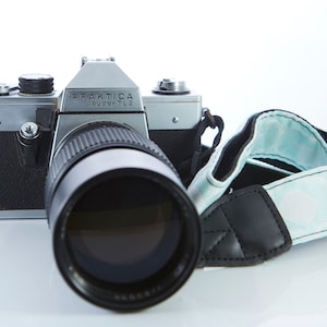 Op de afbeelding: Een zilveren en zwarte Praktica Super TL2 camera met een zwarte lens en een lichtblauwe en zwarte cameraband.