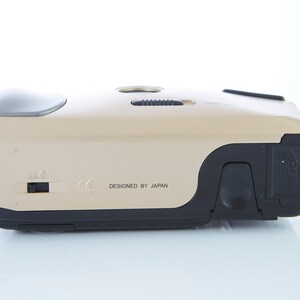 May include: A gold-colored compact camera with black accents. The camera has a lens cover, a flash, and a zoom control. The text "DESIGNED BY JAPAN" is printed on the bottom of the camera.