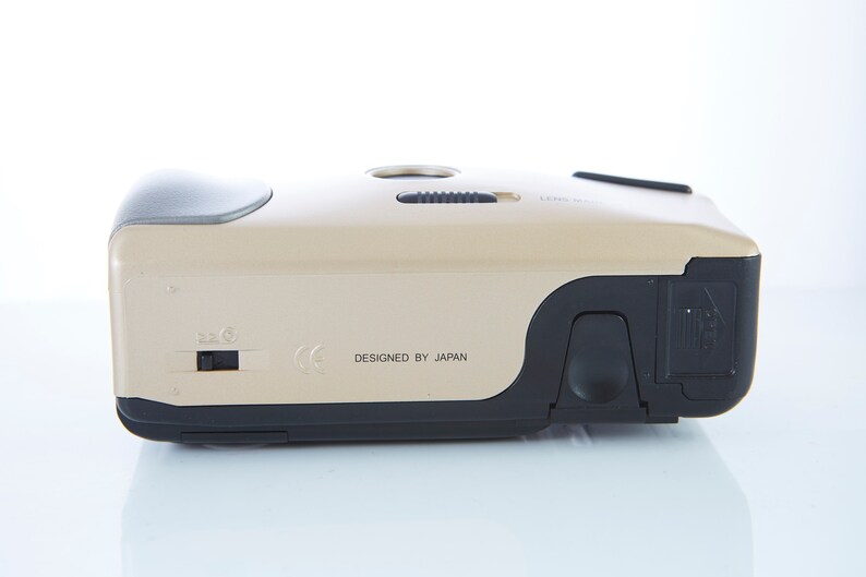 May include: A gold-colored compact camera with black accents. The camera has a lens cover, a flash, and a zoom control. The text "DESIGNED BY JAPAN" is printed on the bottom of the camera.