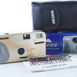 May include: A gold and silver 35mm film camera with a black carrying case and instructions. The camera is labeled "SKINA SK-5550 Motor Drive Auto Flash Lens Made in Japan".
