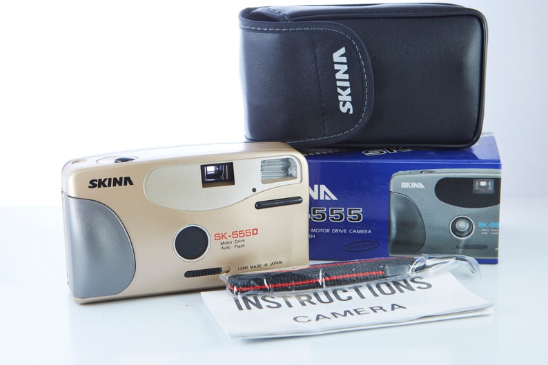 May include: A gold and silver 35mm film camera with a black carrying case and instructions. The camera is labeled "SKINA SK-5550 Motor Drive Auto Flash Lens Made in Japan".