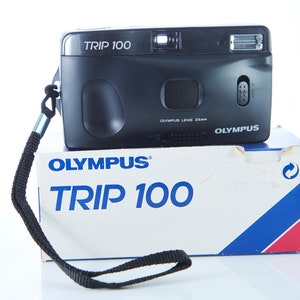May include: A black Olympus Trip 100 film camera with a black strap, sitting on top of its original white box with blue and red accents. The box has the text "Olympus Trip 100" printed on it.