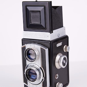 May include: A black and silver Weltaflex camera with a lens that says "Recta 105 5.5" and "Germany". The camera has a built-in flash and a viewfinder.