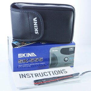 May include: A black camera case with the word "SKINA" printed on it. The case is sitting on top of a blue box with a silver camera on it. The box has the words "SKINA SK-555 35mm COMPACT MOTOR DRIVE CAMERA WITH AUTO FLASH" printed on it. There is also a white instruction booklet with the word "INSTRUCTIONS" printed on it.