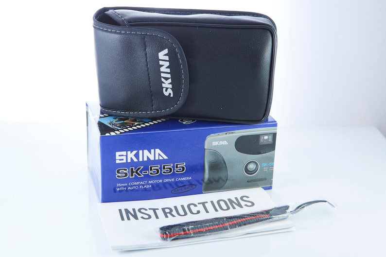May include: A black camera case with the word "SKINA" printed on it. The case is sitting on top of a blue box with a silver camera on it. The box has the words "SKINA SK-555 35mm COMPACT MOTOR DRIVE CAMERA WITH AUTO FLASH" printed on it. There is also a white instruction booklet with the word "INSTRUCTIONS" printed on it.