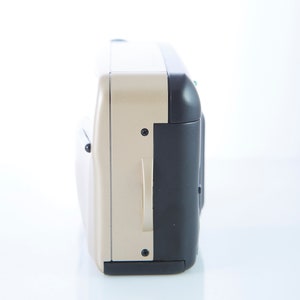 May include: A gold and black compact camera with a built-in flash. The camera has a small LCD screen on the back.