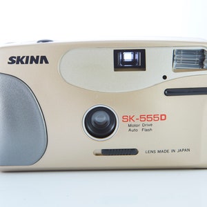 May include: A gold-colored SKINA SK-555D 35mm film camera with a black lens and a built-in flash. The camera has a motor drive and auto flash features. The text "LENS MADE IN JAPAN" is printed on the bottom of the camera.