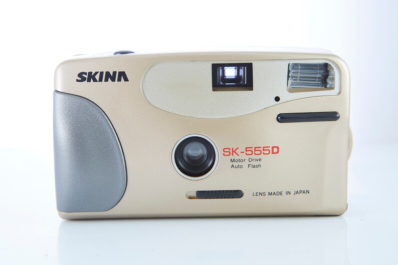 May include: A gold-colored SKINA SK-555D 35mm film camera with a black lens and a built-in flash. The camera has a motor drive and auto flash features. The text "LENS MADE IN JAPAN" is printed on the bottom of the camera.