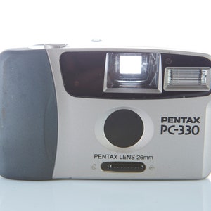 Puede incluir: Una cámara compacta de apuntar y disparar Pentax PC-330 plateada y gris con una lente negra y un flash incorporado. La cámara tiene una correa negra.