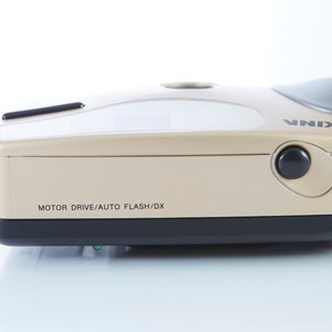May include: A gold-colored camera with a black lens and a black button on the side. The camera has the text "MOTOR DRIVE/AUTO FLASH/DX" and "SKINA" printed on it.