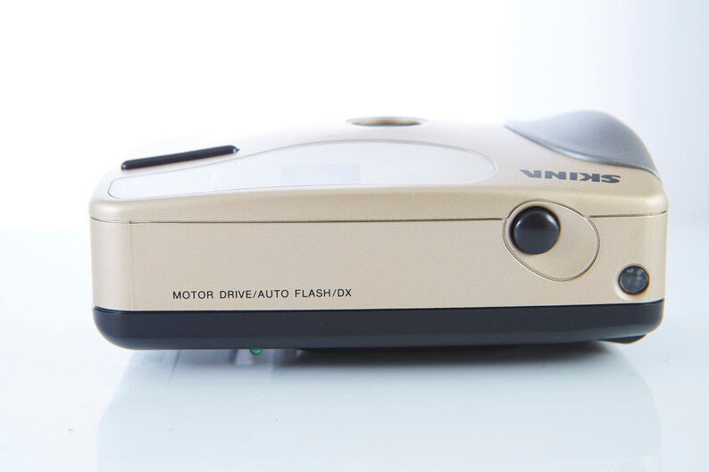 May include: A gold-colored camera with a black lens and a black button on the side. The camera has the text "MOTOR DRIVE/AUTO FLASH/DX" and "SKINA" printed on it.