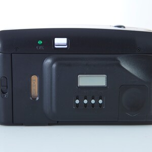 May include: Black compact film camera with a digital display, a small green light, and a silver button. The camera has a built-in flash and a zoom lens.