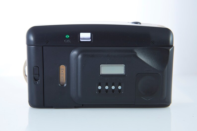 May include: Black compact film camera with a digital display, a small green light, and a silver button. The camera has a built-in flash and a zoom lens.