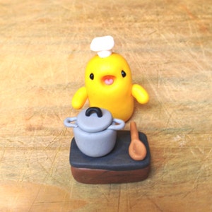 Puede incluir: Una figurita de arcilla amarilla de un chef con un gorro de cocinero blanco, de pie junto a una olla gris sobre una estufa marrón y negra con una cuchara de madera.