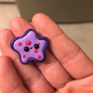 May include: A handcrafted, purple starfish-shaped charm with a smiling face, black eyes, and red spots. The charm has a dark purple border and is held in a hand.