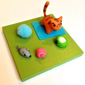 May include: A handmade cat-themed diorama featuring an orange cat with brown stripes, a blue ball, a gray mouse, a pink ball, and a green bowl with a white center. The items are arranged on a green square.