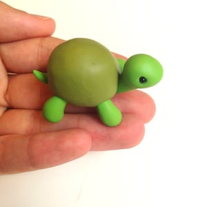 Puede incluir: Una pequeña figura de tortuga verde con caparazón marrón, hecha de arcilla polimérica. La tortuga está sentada en una mano.