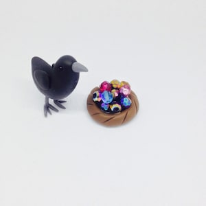 May include: A black bird figurine looking at a small brown clay nest filled with colorful iridescent beads.