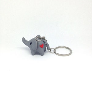 May include: A grey elephant keyring with a red heart on its back. The keyring is attached to a silver key ring.