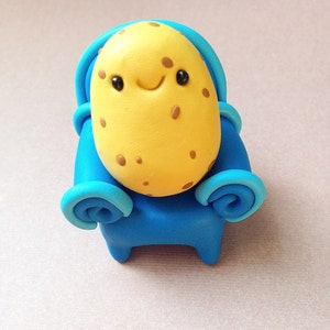 Puede incluir: Figura de arcilla hecha a mano que representa un personaje de patata sonriente. La patata tiene un cuerpo amarillo con manchas marrones, ojos negros y una sonrisa curvada. Lleva un atuendo azul con capucha y detalles de brazos rizados.