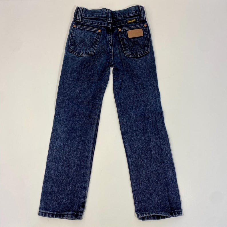 Może przedstawiać: Dżinsy z ciemnego denimu z brązową sk&oacute;rzaną naszywką na tylnej kieszeni z napisem "Wrangler".