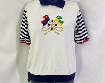 May include: White short-sleeved shirt with a blue and white striped sleeve, a blue collar, and a navy blue band at the bottom. The shirt features an embroidered design of two crossed flags with a rope and a nautical star.