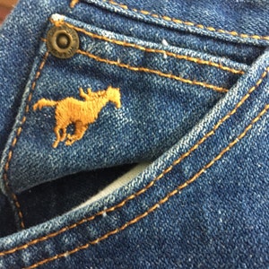 Op de afbeelding: Close-up van een blauwe denimzak met een bruin paardenlogo en een metalen knoop. De zak heeft een dubbele rij stiksels rond de rand.
