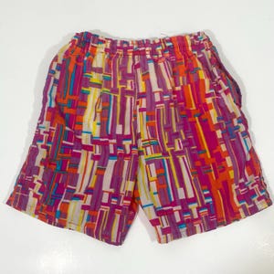 May include: Colorful shorts with an abstract geometric pattern. The shorts feature a vibrant mix of colors, including purple, orange, yellow, and white. The elastic waistband is visible at the top. The shorts are made of a lightweight fabric.