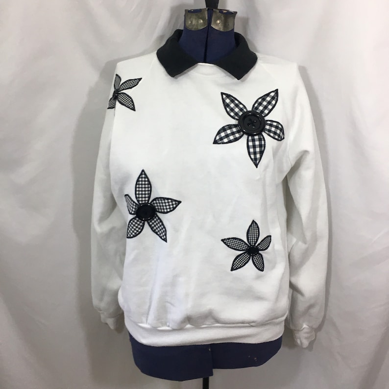 Puede incluir: Sudadera blanca con cuello negro y cuatro flores de vichy en blanco y negro con botones negros en el centro.