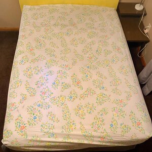 Pode incluir: Um lençol de cama branco com um padrão floral em tons de rosa, azul, amarelo e verde. O lençol é feito de um tecido macio e confortável e tem um brilho sutil.