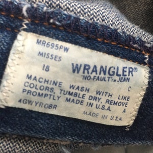 Op de afbeelding: Een wit label met zwarte tekst op een blauwe denim achtergrond. Het label luidt "MR695PW Misses 18 Wrangler No-Fault Jean C Machine Wash With Like Colors, Tumble Dry, Remove Promptly Made in U.S.A. 4GWYROBR Made in U.S.A. A"