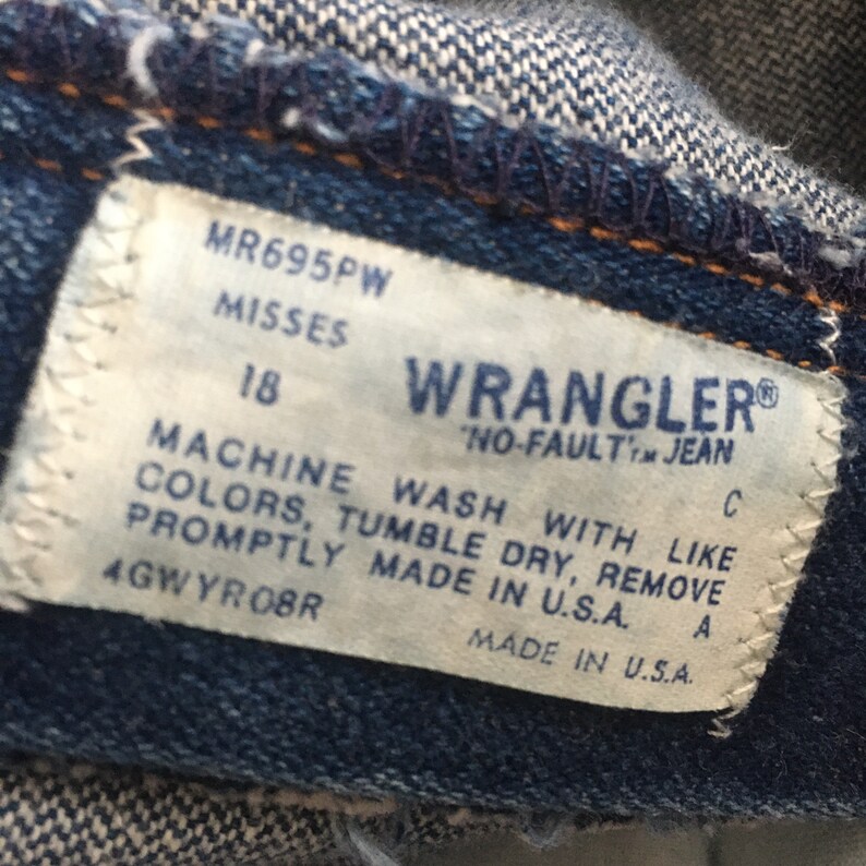 Op de afbeelding: Een wit label met zwarte tekst op een blauwe denim achtergrond. Het label luidt "MR695PW Misses 18 Wrangler No-Fault Jean C Machine Wash With Like Colors, Tumble Dry, Remove Promptly Made in U.S.A. 4GWYROBR Made in U.S.A. A"