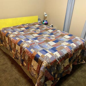 May include: A patchwork quilt in shades of blue, red, brown, and cream covers a bed with a yellow headboard. The quilt features a variety of patterned fabric squares sewn together. The bed is set against a neutral-colored wall.