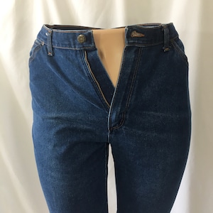 Op de afbeelding: Een donkerblauwe spijkerbroek met een ritsvlieg en een knoopsluiting. De broek is opengeknoopt en de rits is open.