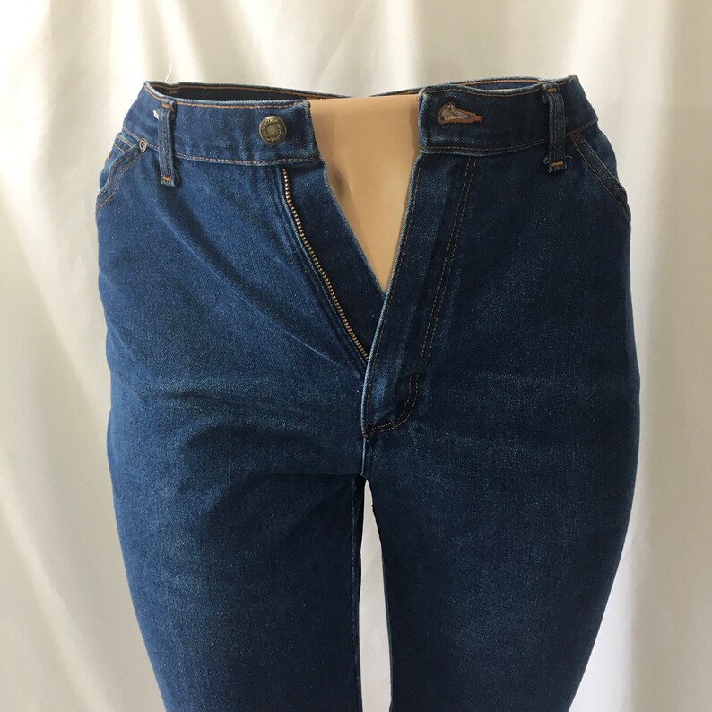 Op de afbeelding: Een donkerblauwe spijkerbroek met een ritsvlieg en een knoopsluiting. De broek is opengeknoopt en de rits is open.