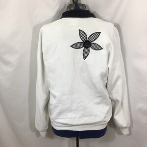 Puede incluir: Sudadera blanca con una aplicaci&oacute;n de flor a cuadros negros y blancos en la espalda. La flor tiene un bot&oacute;n negro en el centro.