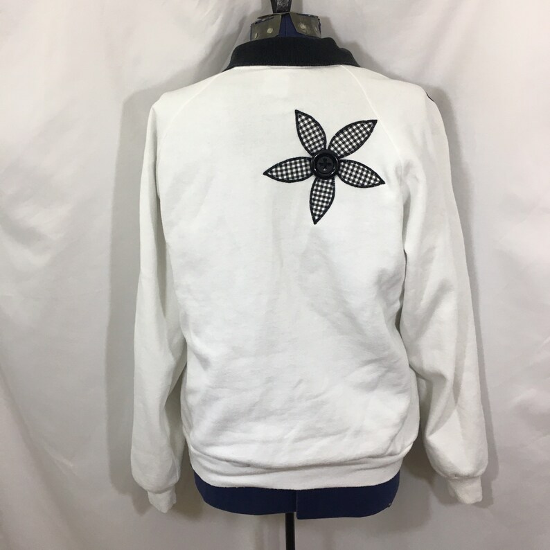 Puede incluir: Sudadera blanca con una aplicaci&oacute;n de flor a cuadros negros y blancos en la espalda. La flor tiene un bot&oacute;n negro en el centro.