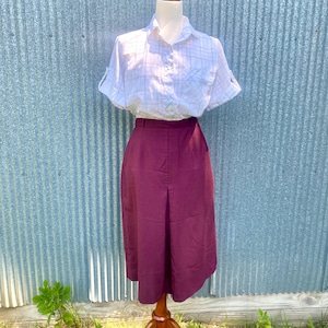 May include: A white short-sleeved button-down shirt with a light blue and grey plaid pattern paired with a solid deep burgundy midi-length skirt featuring a central front pleat, displayed on a mannequin against a corrugated metal background.