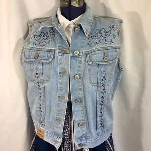 May include: A light blue denim vest with embroidered birds and hearts. The vest has a button-up front and two pockets.