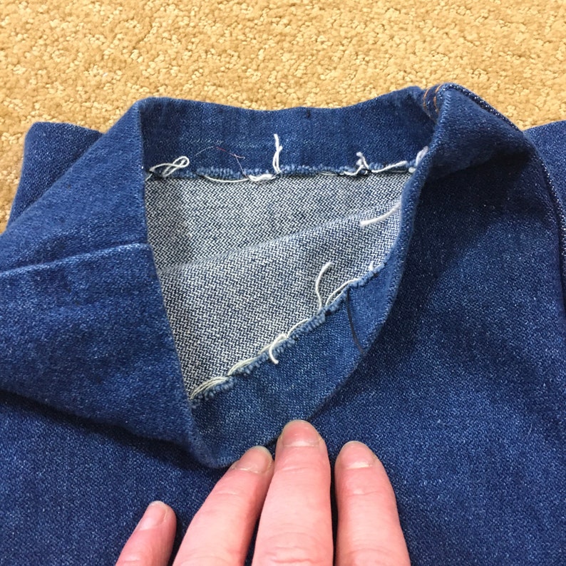 Op de afbeelding: Close-up van de binnenkant van een blauwe denim jackenkraag, die de rauwe rand van de naad en de stiksels laat zien.