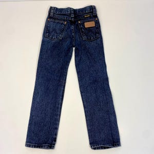 Może przedstawiać: Dżinsy z ciemnego denimu z brązową sk&oacute;rzaną naszywką na tylnej kieszeni z napisem "Wrangler".
