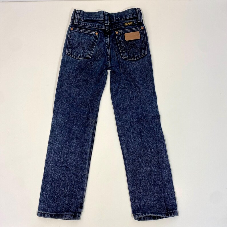 Może przedstawiać: Dżinsy z ciemnego denimu z brązową sk&oacute;rzaną naszywką na tylnej kieszeni z napisem "Wrangler".
