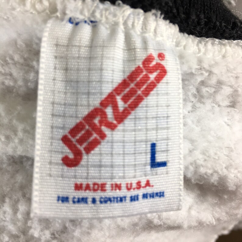 Puede incluir: Etiqueta blanca con texto rojo que dice "JERZEES" y una "L" azul para el tama&ntilde;o. La etiqueta tambi&eacute;n dice "MADE IN U.S.A." y "FOR CARE & CONTENT SEE REVERSE".
