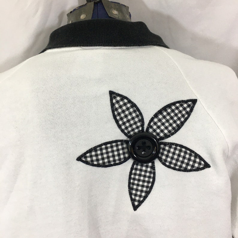 Puede incluir: Sudadera blanca con una aplicaci&oacute;n de flor de vichy en blanco y negro y un bot&oacute;n negro en el centro.