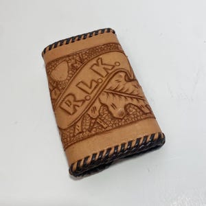 May include: A brown leather wallet with a black stitched border. The wallet has a tooled design with the word "RLK" embossed on the front.