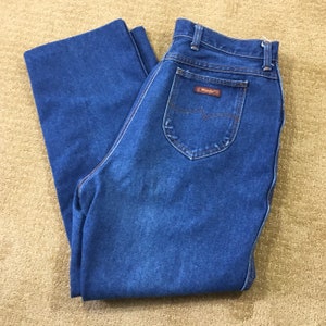 Op de afbeelding: Een paar blauwe denim jeans met een bruin leren patch op de achterzak met de tekst "Wrangler".