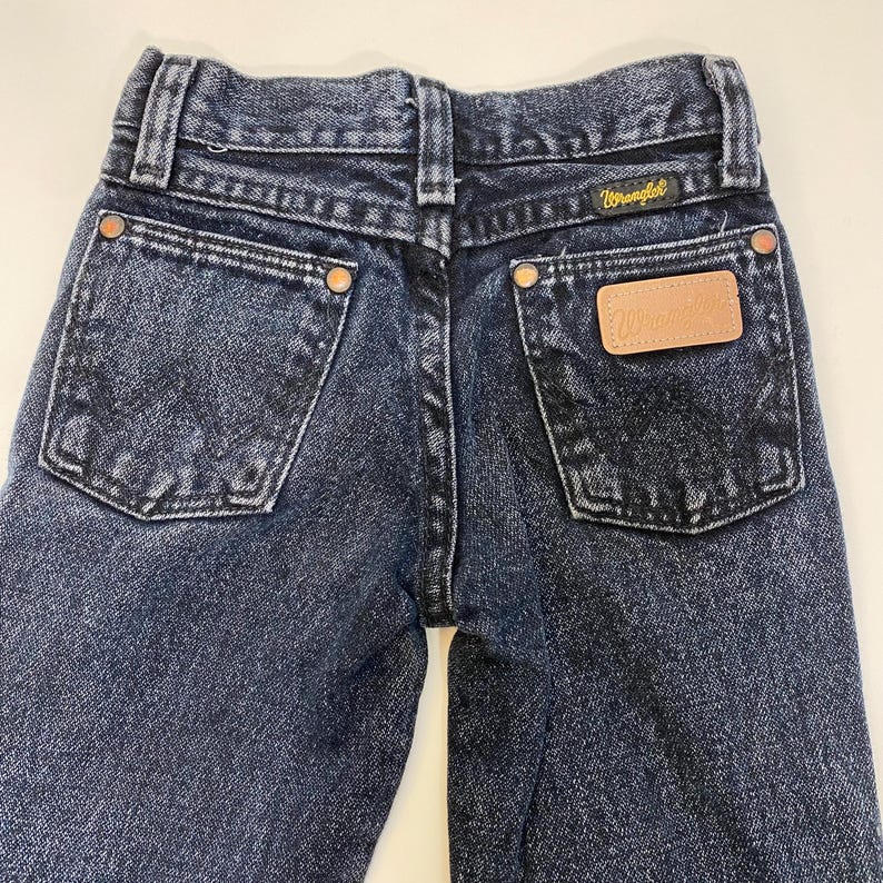 Może przedstawiać: Dżinsy z ciemnego denimu z sk&oacute;rzaną naszywką na tylnej kieszeni z napisem "Wrangler".